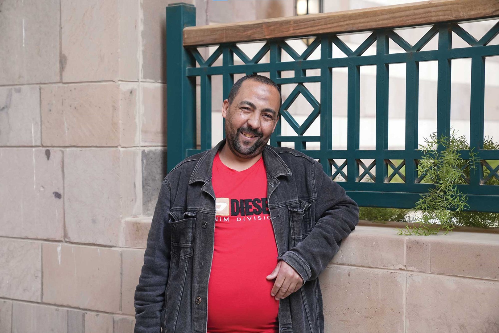 Mohamed Salah, known as Tata, stands smiling outdoors on AUC&rsquo;s campus, leaning against a low wall, wearing a red T-shirt and dark jacket.