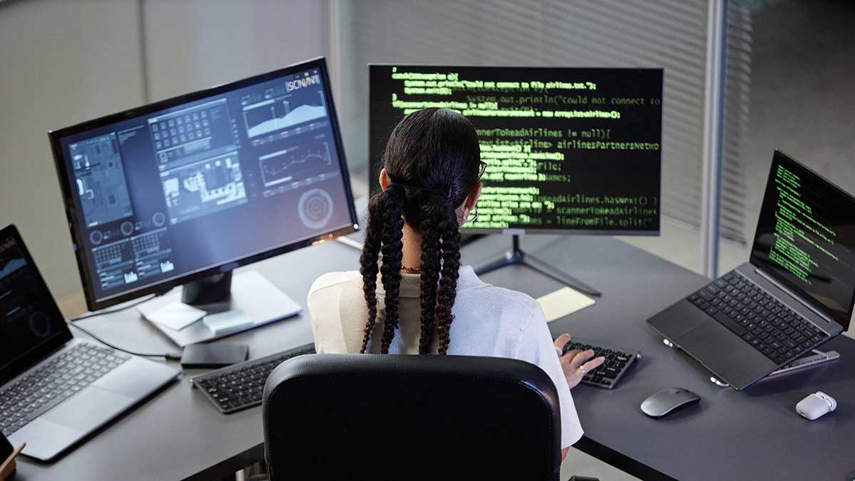 A woman codes on two monitor screens and a laptop