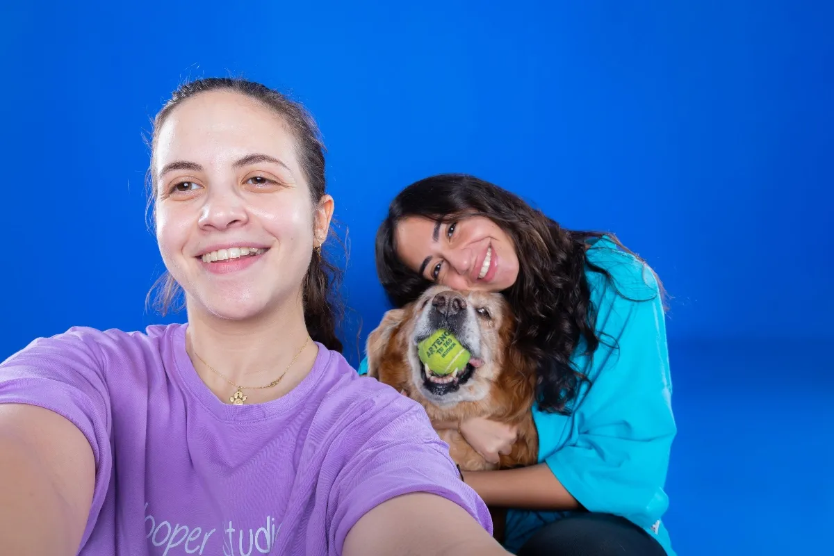 two women take a selfie with a golden retriever holding a tennis ball in her mouth