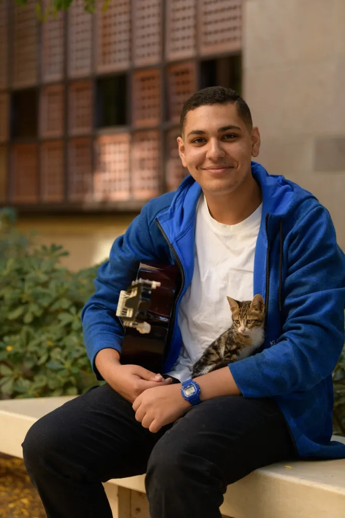 man wearing a white t shirt and a blue jacket sits outside on a bench among greenery with a cat on his lap and a guitar tucked under his right arm