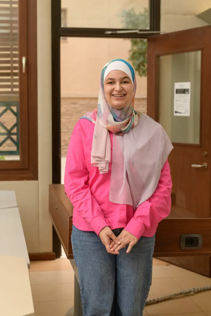 Woman wearing jeans, a bright pink shirt, and a white, blue, and pink headscarf leans against a desk in a classroom, smiling