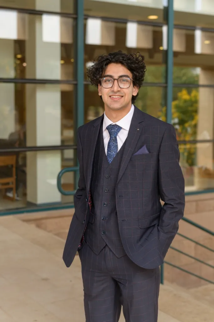 Man wearing suit and glasses stands smiling in front of glass windows