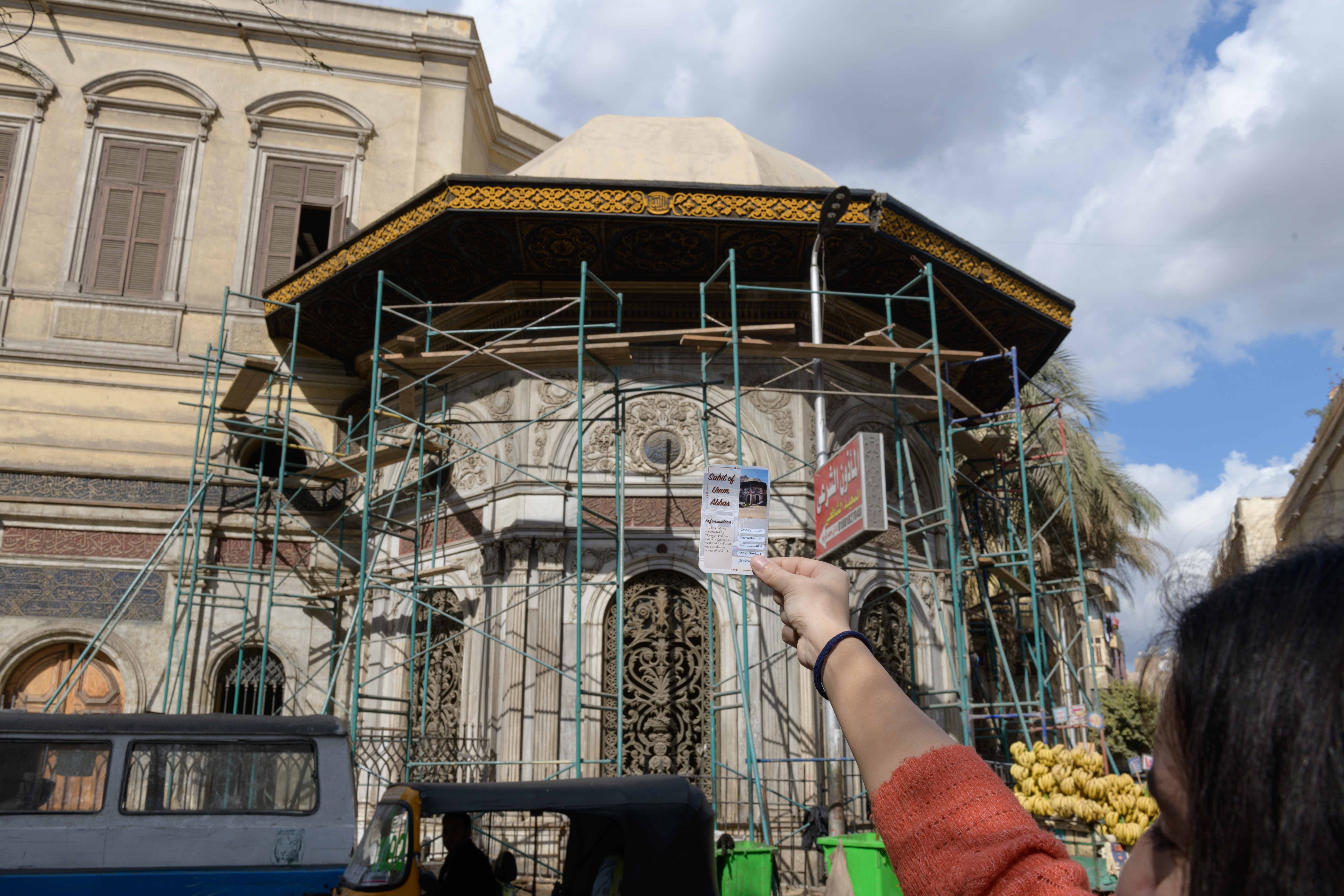 hand holds up a card depicting a sabil in front of the sabil itself