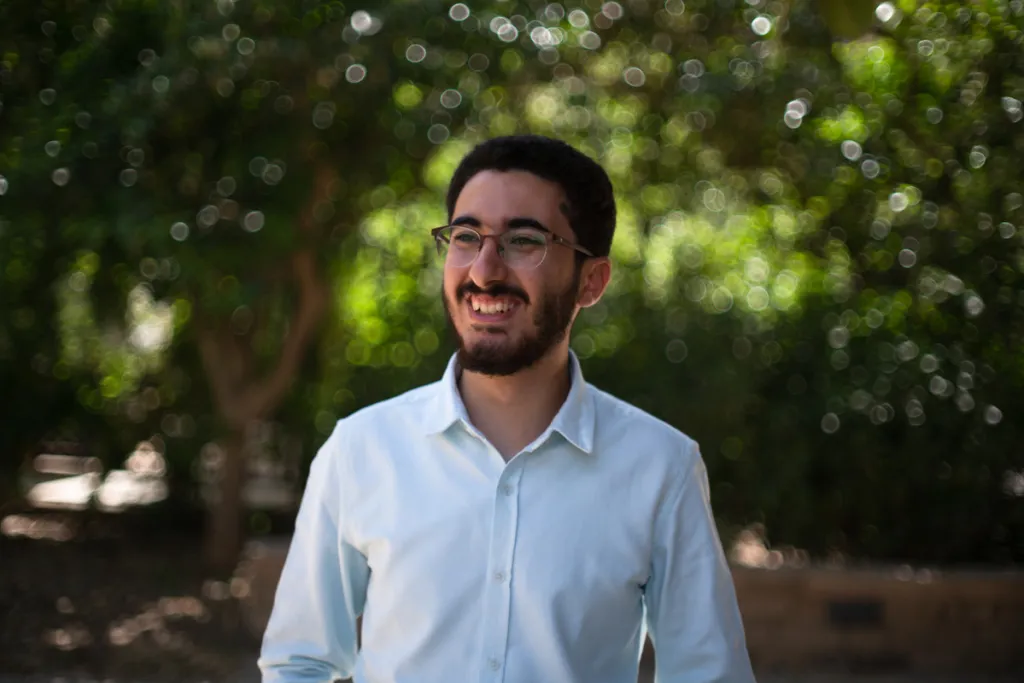 Man wearing a blue button-up shirt smiles standing in front of trees