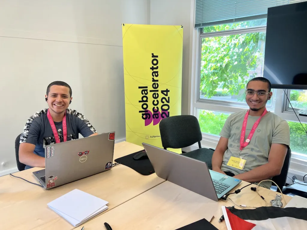 two men sit at a desk on their laptops looking at the camera and smiling
