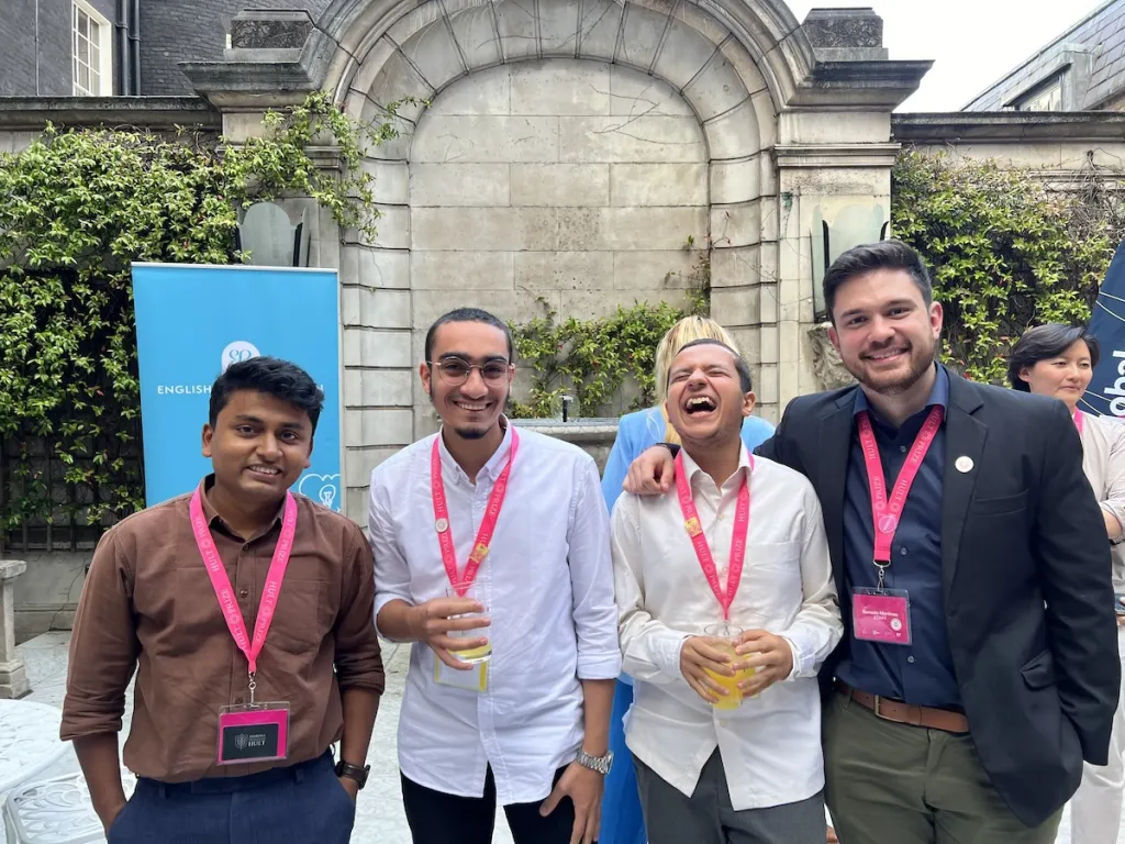 four men stand outside in a courtyard smiling, one is laughing