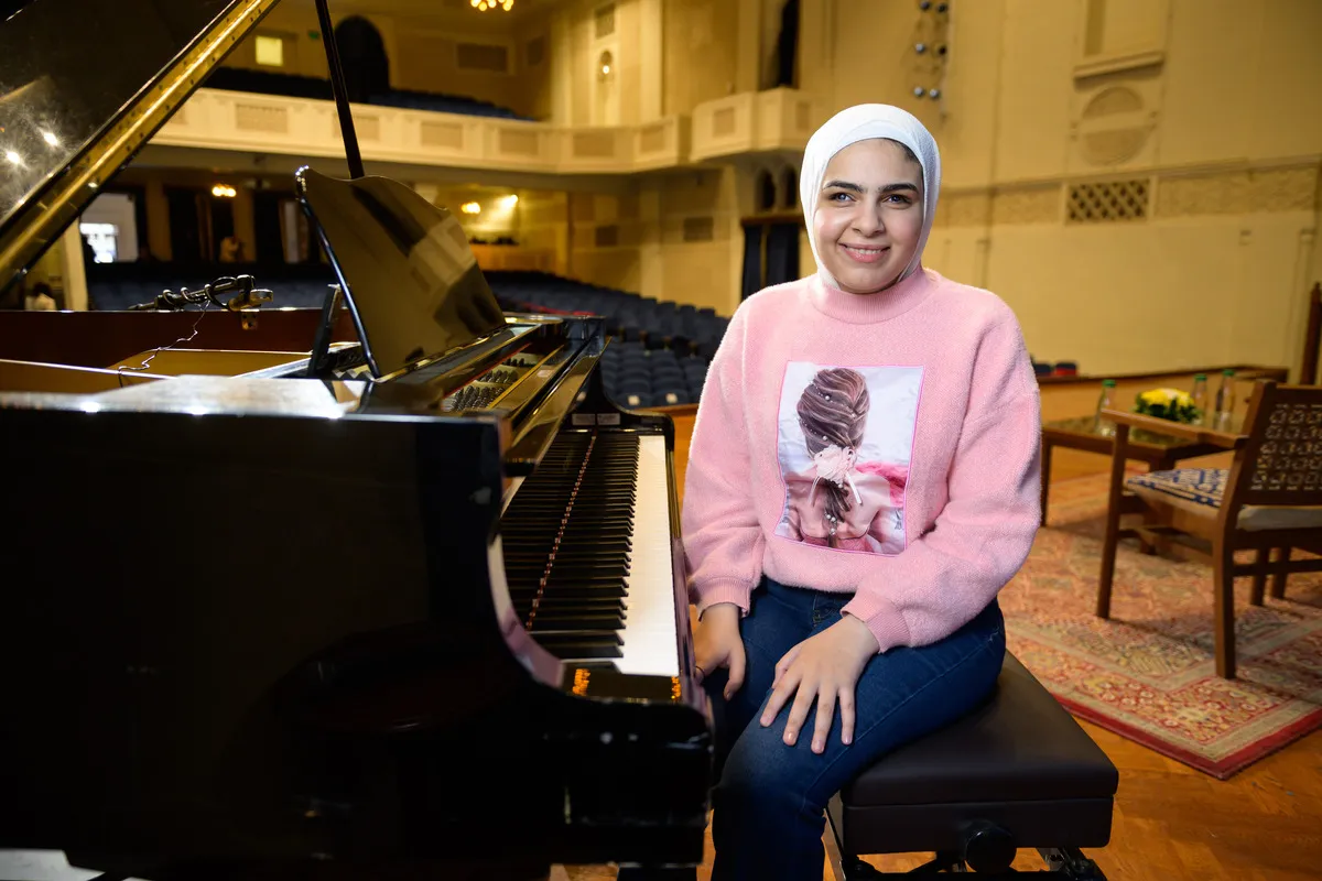woman sits on a piano bench in front of a piano on stage in an auditorium. She is facing the camera and smiling