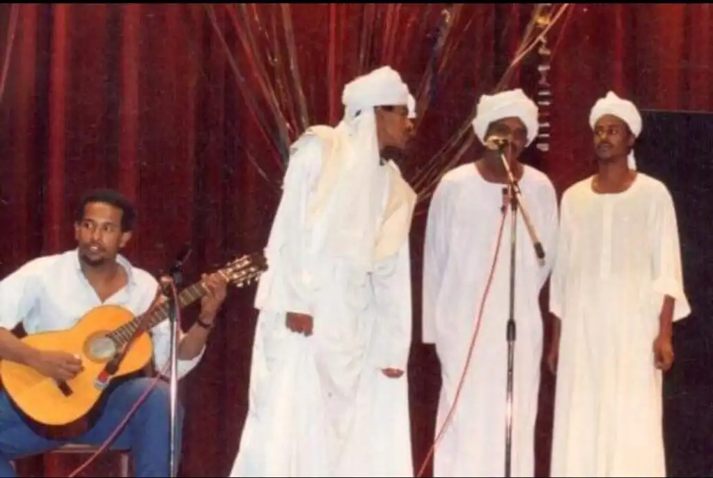 men wearing white stand on stage singing into a microphone while another man sits behind them playing guitar