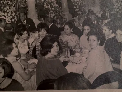a group of smiling women at a dining table, dressed up, in black and white.
