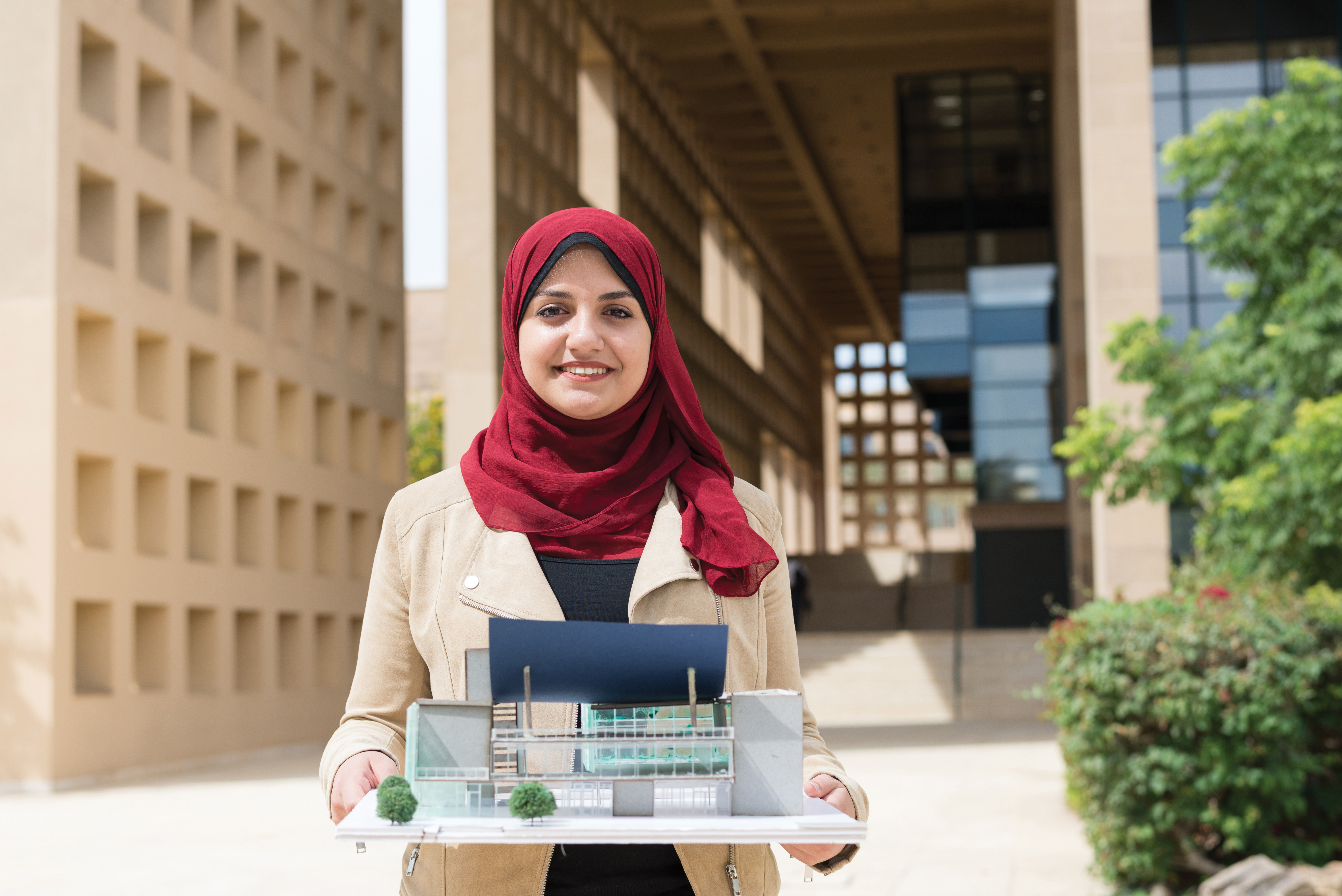 A female student in a red hijab holds her architecture project, a model home, and smiles straight ahead.
