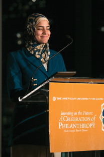 A woman in a teal and tan hijab smiles at a podium
